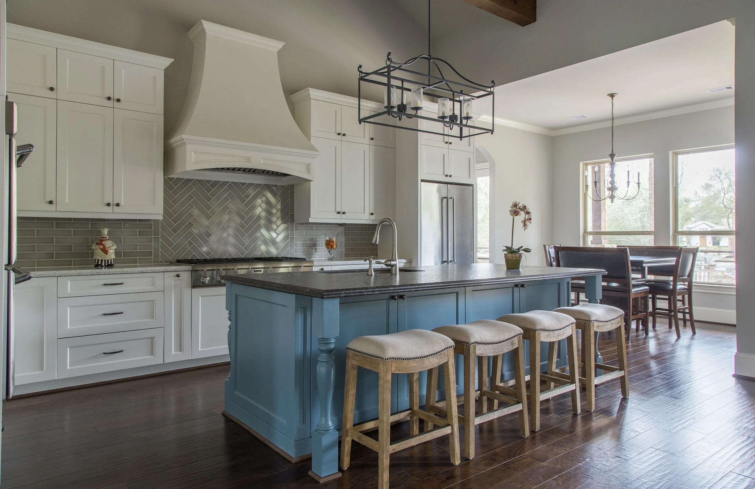 a modern kitchen remodeling consultation in a stylish urban office, showcasing blueprints and material samples on a sleek conference table, with an expert advisor engaged in discussion with a client. A stylish kitchen with white cabinetry, a dramatic white range hood, and a blue island topped with a dark countertop. Four cushioned wooden bar stools line the island, while herringbone-patterned backsplash tiles and stainless steel appliances add elegance. The space also features dark wood floors, a modern chandelier above the island, and a dining area with large windows in the background.
